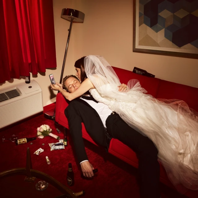 Newlyweds lounge on a red sofa, the bride holding a phone, in a motel room littered with champagne bottles and a spilled bouquet of flowers.