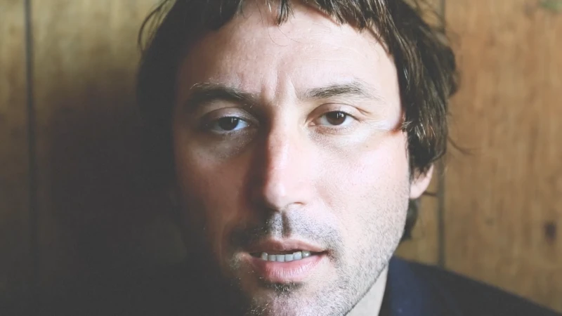 Close-up portrait of a man with dark hair and stubble looking directly forward against a wooden wall background.