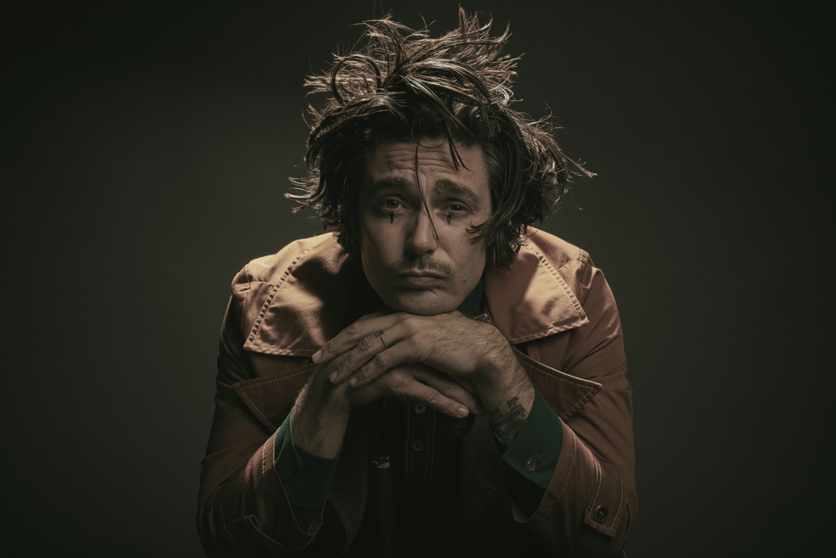 Portrait of an adult male with tousled hair and face makeup, resting his chin on interlocked fingers, wearing a rust-colored jacket in a studio.