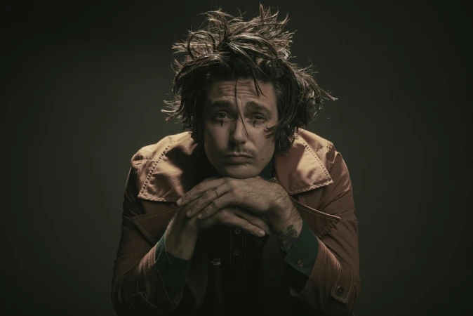 Portrait of an adult male with tousled hair and face makeup, resting his chin on interlocked fingers, wearing a rust-colored jacket in a studio.