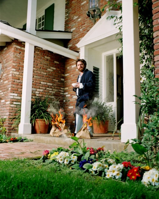 A man in pajamas on a porch holding a mug, with flaming paper bags nearby and flowers in the front yard of a brick house.