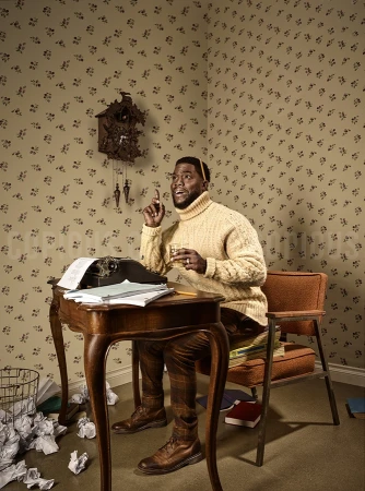 Man sits at desk with typewriter in retro wallpaper room, raises finger, holds glass. Neutral tone, vintage feel. Adult male indoors.