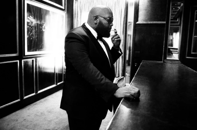 Black and white portrait of a bearded man in a suit and sunglasses smoking indoors near a bar counter by Pamela Littky