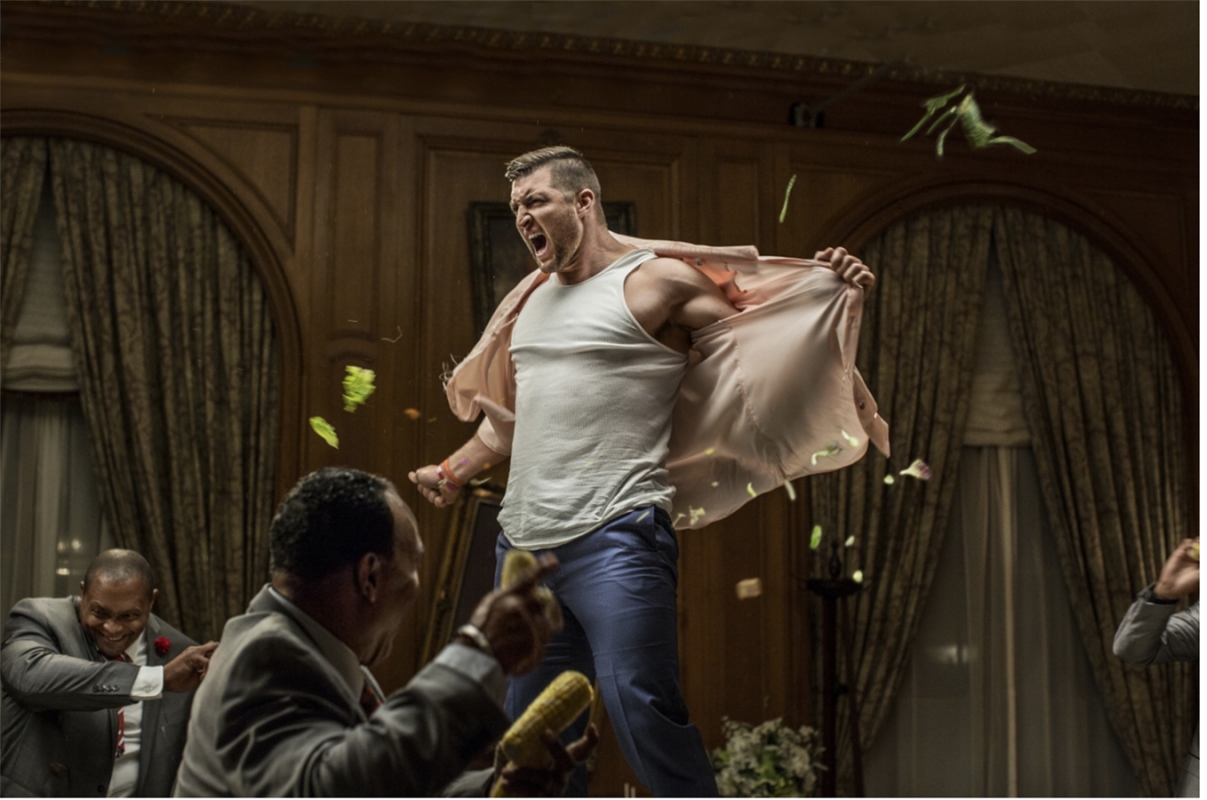 Man in tank top and dress shirt, angry expression, throwing corn cobs surrounded by men in suits in an ornate dining room setting.