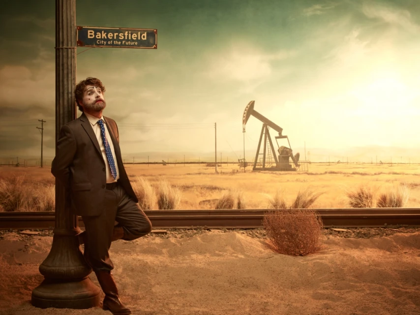 Clown in a suit leans against a pole under a Bakersfield sign with an oil pump in the distance on a golden, hazy day.