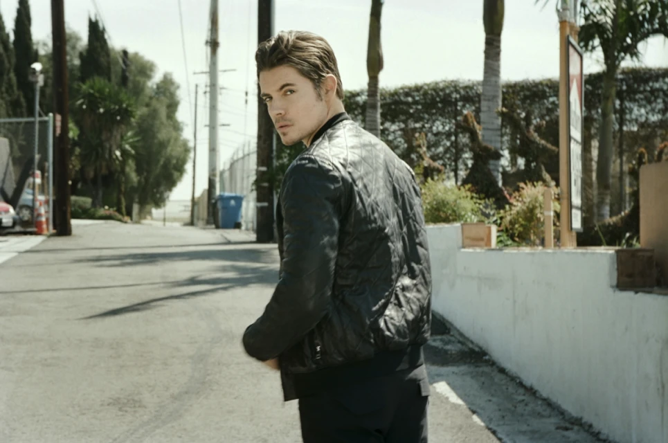 Man in a black jacket glances over his shoulder while walking down a road lined with trees and buildings under a bright sky.
