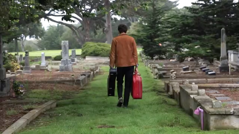 A man walks away from the viewer in a cemetery carrying a guitar case and a red suitcase on a slightly overcast day.