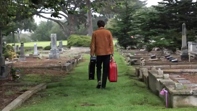 A man walks away from the viewer in a cemetery carrying a guitar case and a red suitcase on a slightly overcast day.