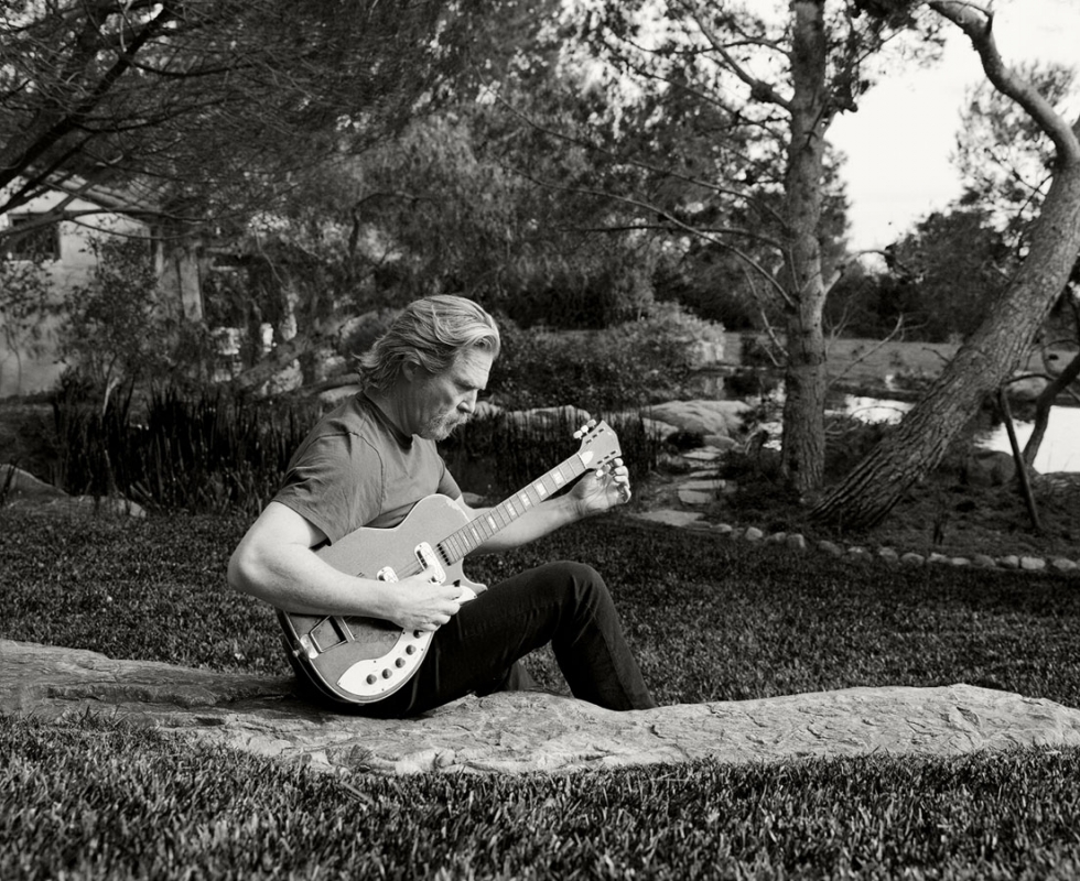 Adult male playing electric guitar while sitting on a rock outdoors on a lawn surrounded by trees in black and white. 