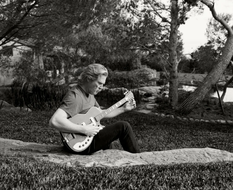 Adult male playing electric guitar while sitting on a rock outdoors on a lawn surrounded by trees in black and white. 