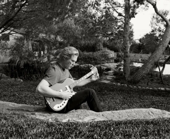 Adult male playing electric guitar while sitting on a rock outdoors on a lawn surrounded by trees in black and white. 
