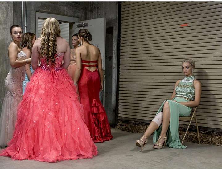 Several young women in formal gowns gather in a backstage area with one seated and pensive, lit by overhead fluorescent lighting.