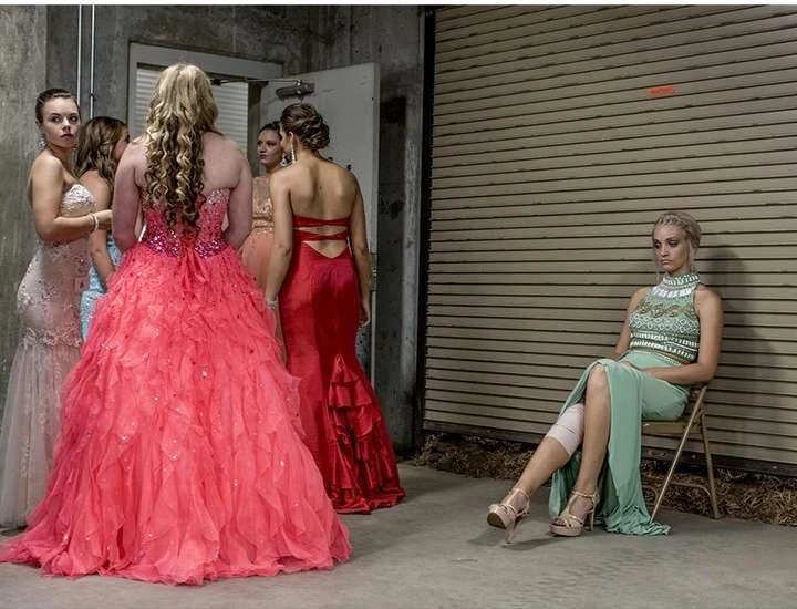 Several young women in formal gowns gather in a backstage area with one seated and pensive, lit by overhead fluorescent lighting.