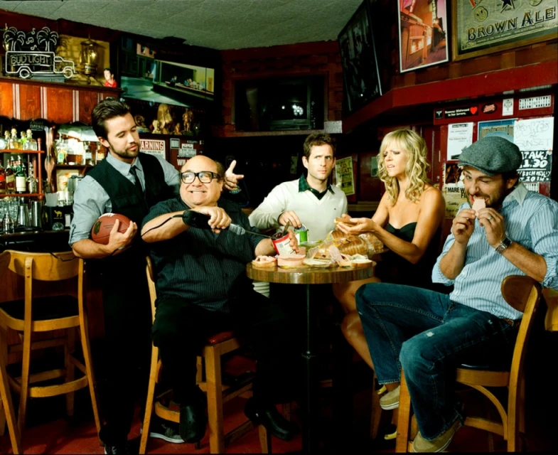 Five people gathered at a bar table with food and drinks, including Danny DeVito. The scene is warmly lit, capturing a casual, social moment.