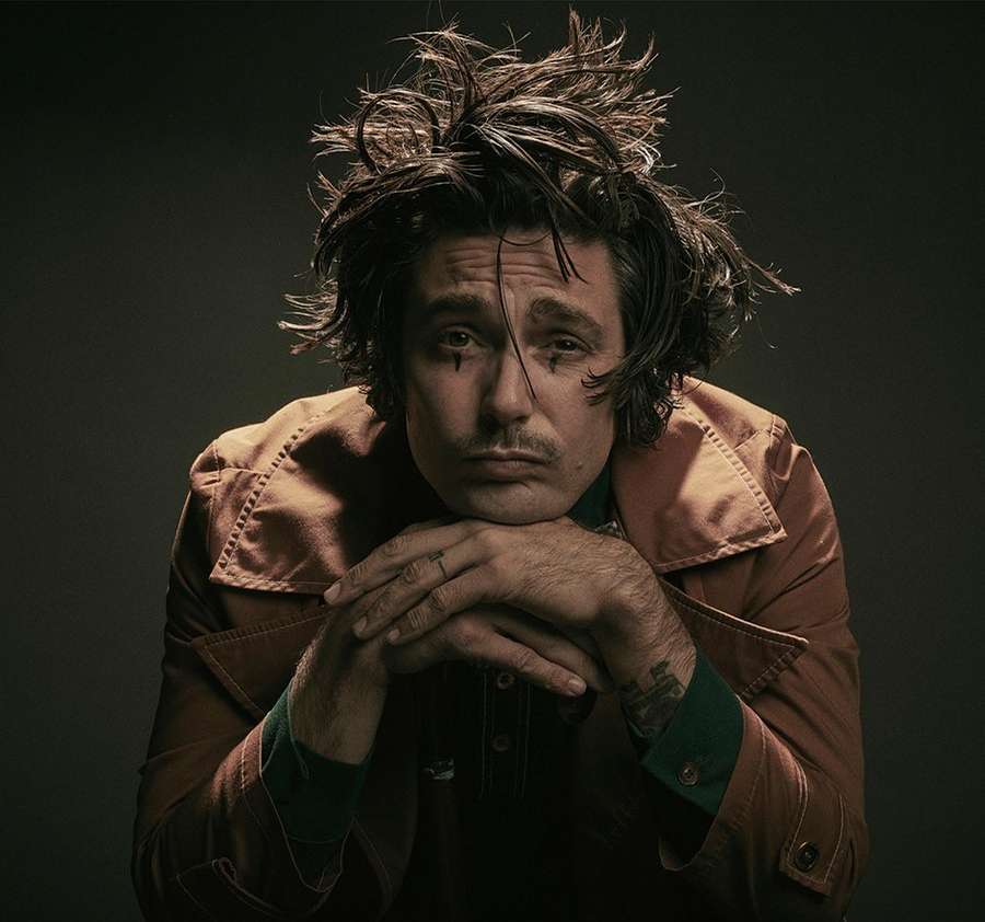 Portrait of a dark-haired adult male with facial markings and messy hair, wearing a brown jacket in a photo studio setting.