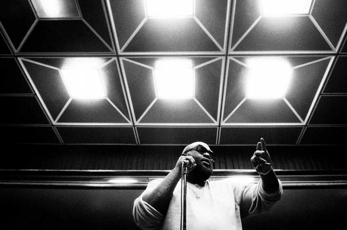 Monochrome shot of a singer holding a microphone and raising a finger under ceiling lights, capturing a musical moment in performance.