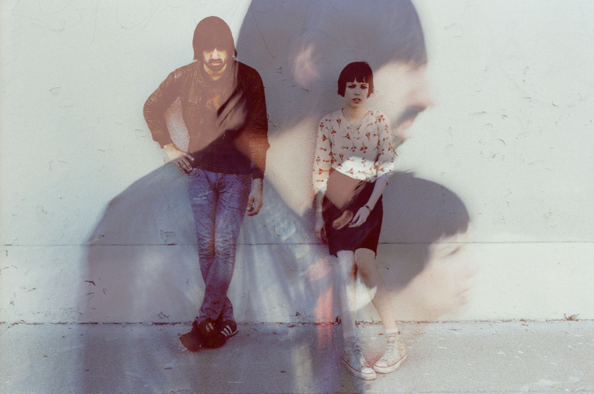A cool duo stands against a textured wall with double exposure effect, casting a shadow of a face; both are dressed in stylish, casual attire.