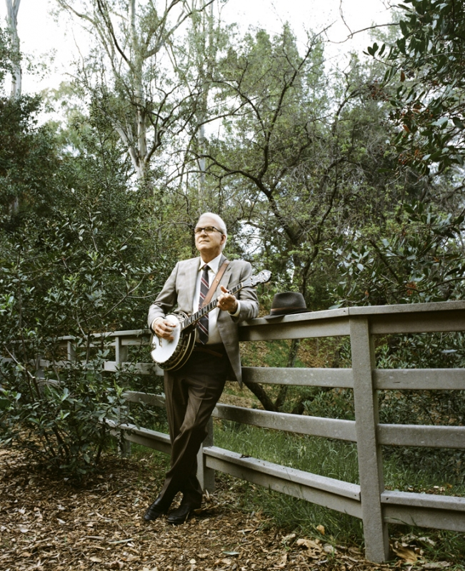 Steve Martin leaning against a wooden fence in a wooded area playing a banjo, wearing a grey suit and tie.