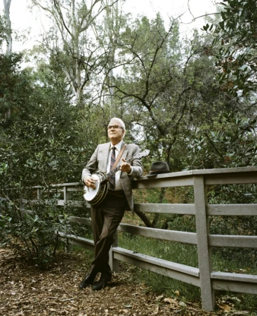 Steve Martin leaning against a wooden fence in a wooded area playing a banjo, wearing a grey suit and tie.