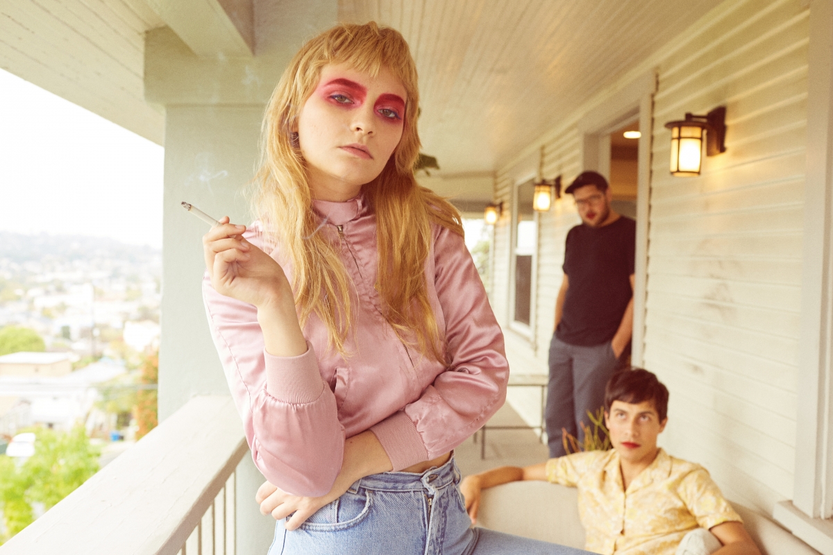 Stylish woman with bold eye makeup smoking on a porch with two men in the background, capturing a vintage or indie aesthetic.