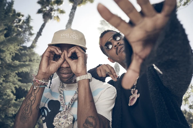 Two men posing outdoors against palm trees, one wearing a baseball cap and making glasses shape with hands.