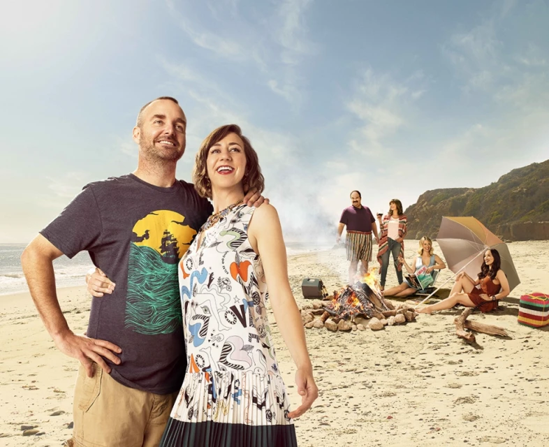 A couple embraces on a sandy beach with friends gathered around a bonfire, set against a backdrop of blue sky and ocean.