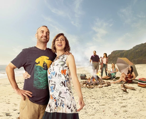 A couple embraces on a sandy beach with friends gathered around a bonfire, set against a backdrop of blue sky and ocean.