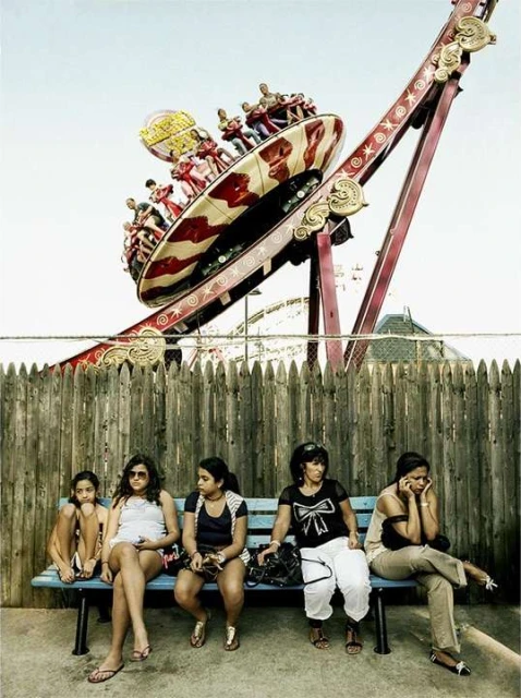 People sit on a bench in front of a fence at an amusement park, with a spinning ride full of people in the background against a blue sky.