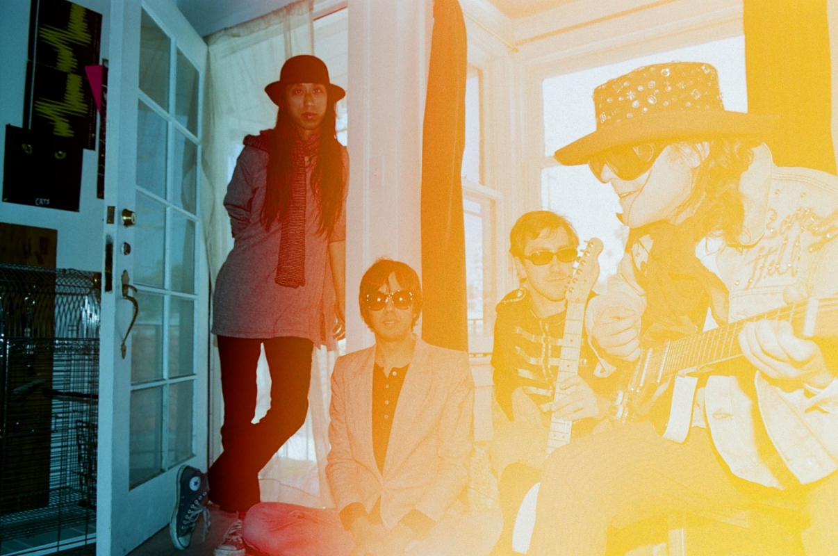 Group of four musicians posing in an interior room with a bright light leak across the right side of the frame.
