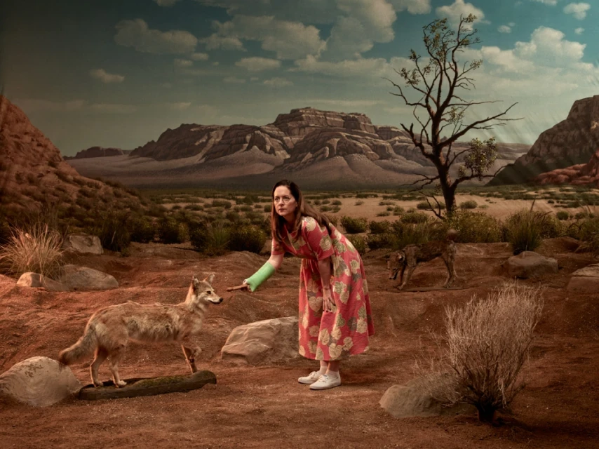 Woman in floral dress stands in a desert landscape with two canids. A painted mountain range and blue sky are in the background.