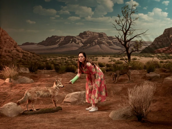 Woman in floral dress stands in a desert landscape with two canids. A painted mountain range and blue sky are in the background.
