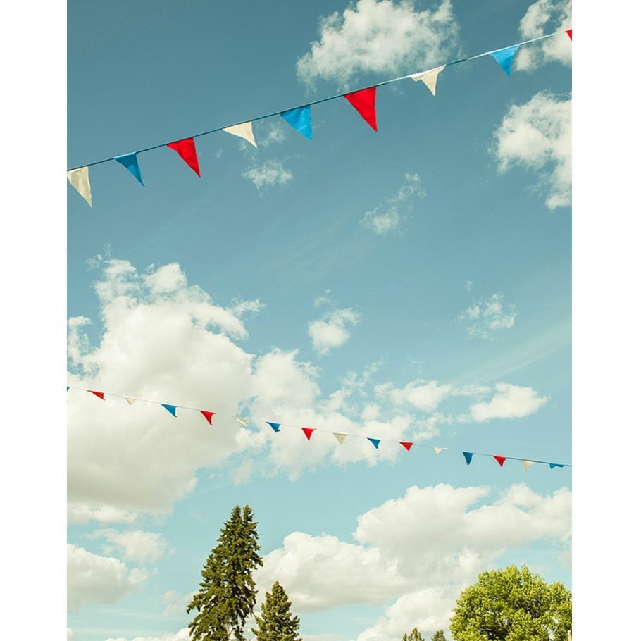 Colorful triangular pennant banners strung against a blue sky with clouds above green trees, creating a festive atmosphere.