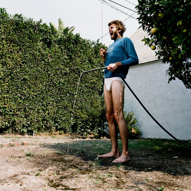 A man in briefs stands on a lawn, shouting and watering the grass with a hose, with a hedge, tree, and house in the background.