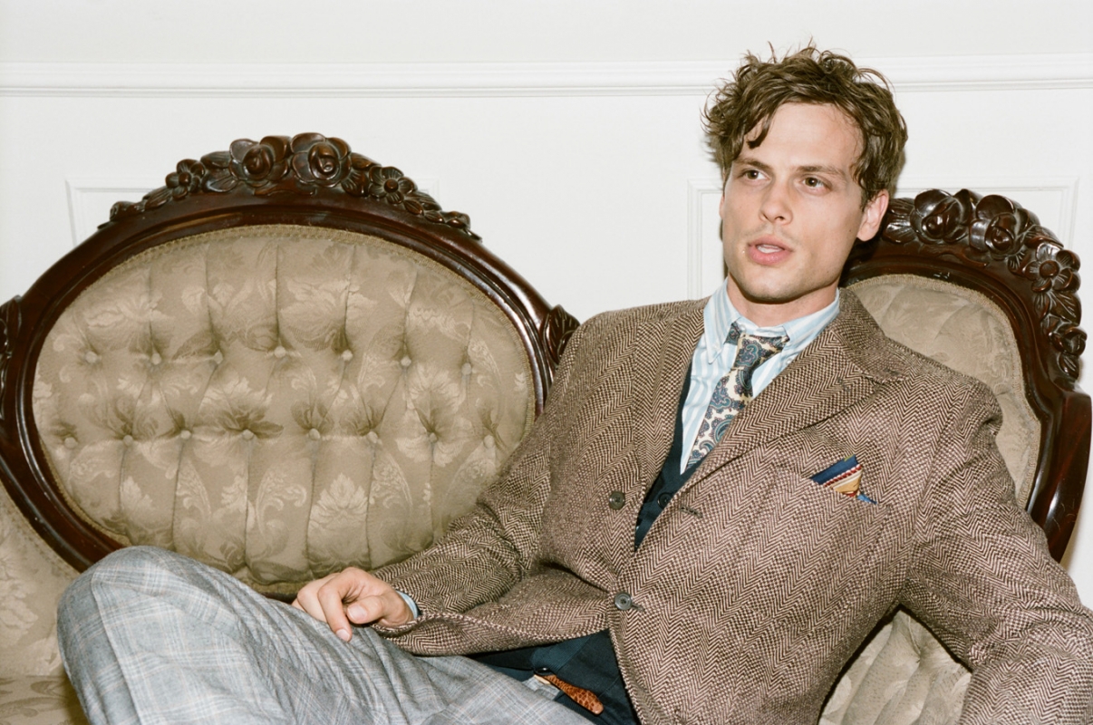 Matthew Gray Gubler poses on a vintage sofa in a herringbone jacket and patterned tie, set against a neutral wall indoors.