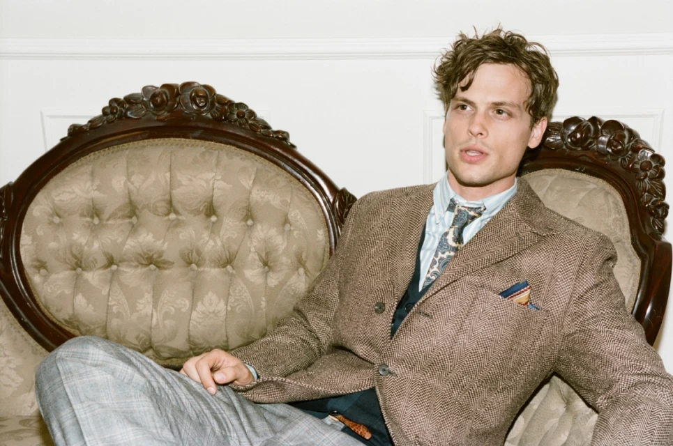 Matthew Gray Gubler poses on a vintage sofa in a herringbone jacket and patterned tie, set against a neutral wall indoors.