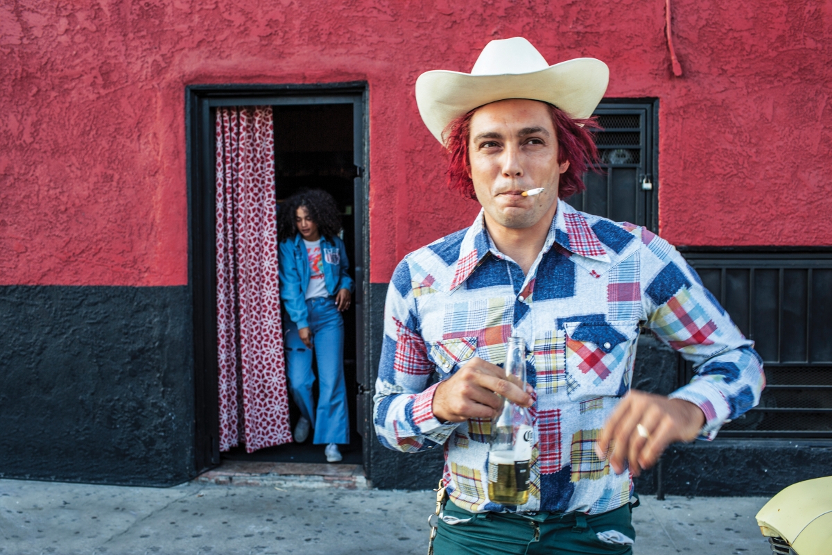 Man in cowboy hat holds beer outside red building, woman in doorway. Bright colors, casual style. Editorial fashion shot. Lifestyle image.