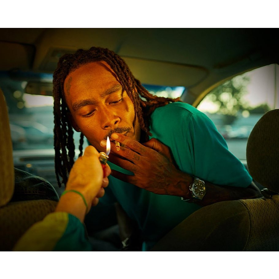 Man with dreadlocks lights a cigarette in car interior. Arm extends lighter. Features teal shirt, watch, vehicle details, subdued lighting.