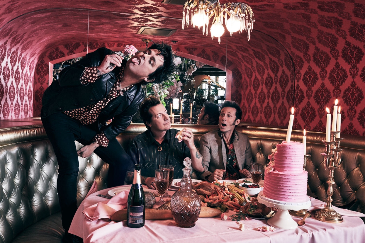 Three adult males pose around a pink-frosted layer cake, sitting at a table inside a restaurant booth with red patterned walls.