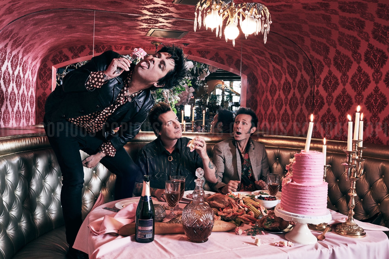 Three adult males gather around a pink-covered table laden with food, a pink cake, and champagne in an ornate, rose-toned room.