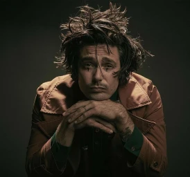 Man with messy dark hair and a mustache resting his chin on his hands, wearing a brown jacket over a dark green shirt.