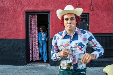 Man wearing a white cowboy hat and patchwork shirt holding a beer outside a red building by Pamela Littky