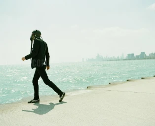 Man wearing a dark suit and helmet walking along a concrete pier next to the water with a distant city skyline