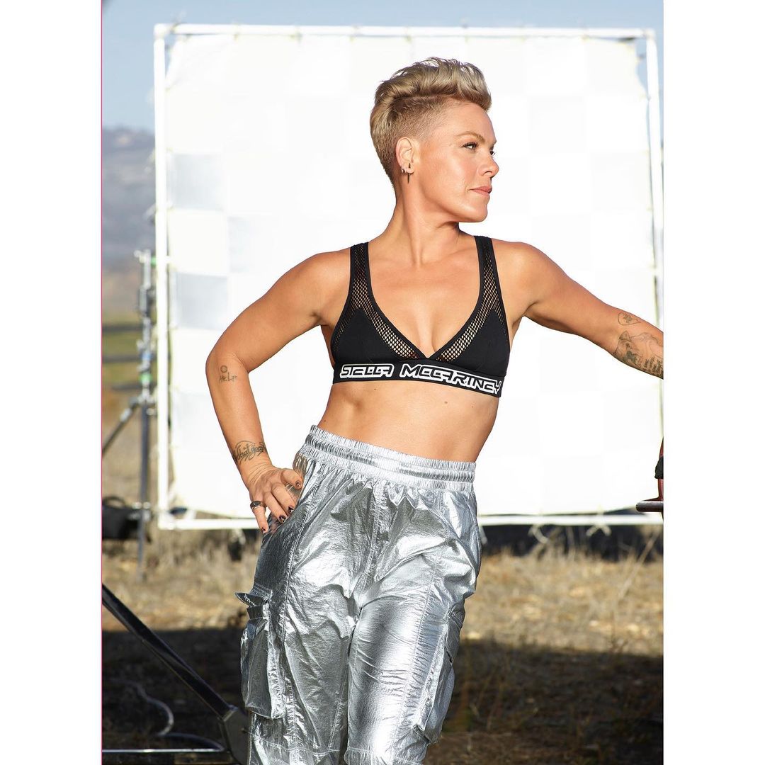 Woman with a pixie haircut poses in a black sports bra and shiny silver pants outdoors against a blurred background and photography equipment.