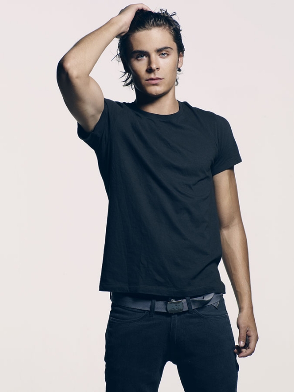 Portrait of a male model with dark hair, wearing a black t-shirt and jeans, posing with his hand on his head against a white background.