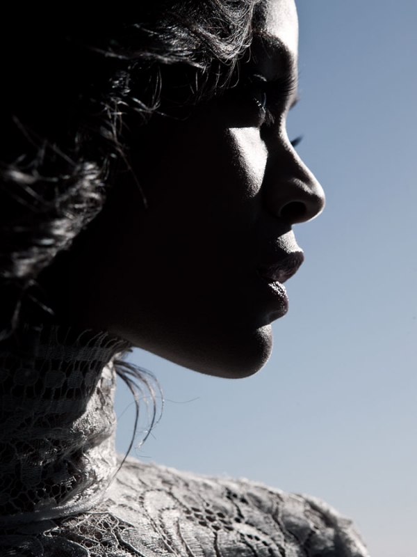 Profile portrait of a woman with dark hair, wearing a lace top, against a gradient blue sky backdrop in bright, diffused lighting.