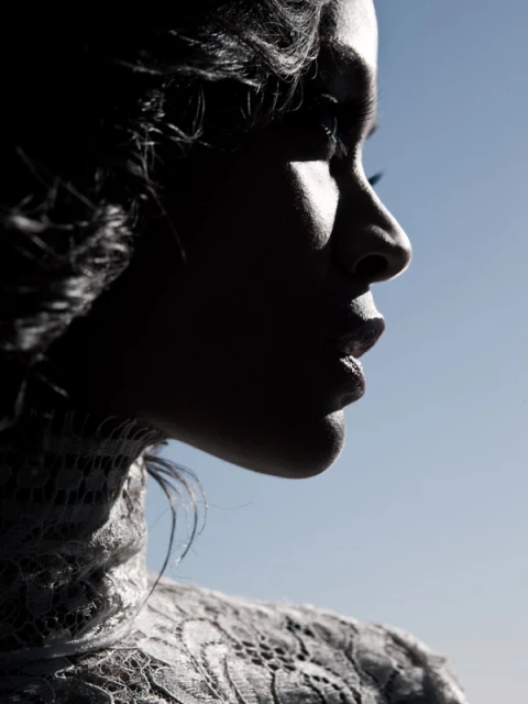Profile portrait of a woman with dark hair, wearing a lace top, against a gradient blue sky backdrop in bright, diffused lighting.