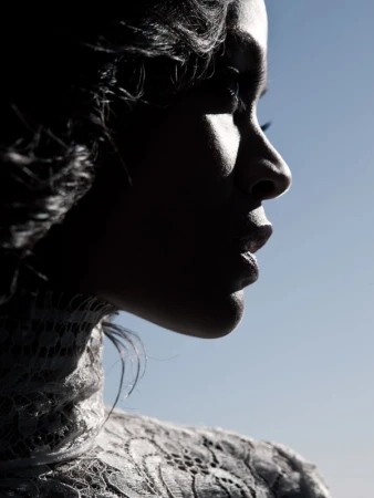 Profile portrait of a woman with dark hair, wearing a lace top, against a gradient blue sky backdrop in bright, diffused lighting.