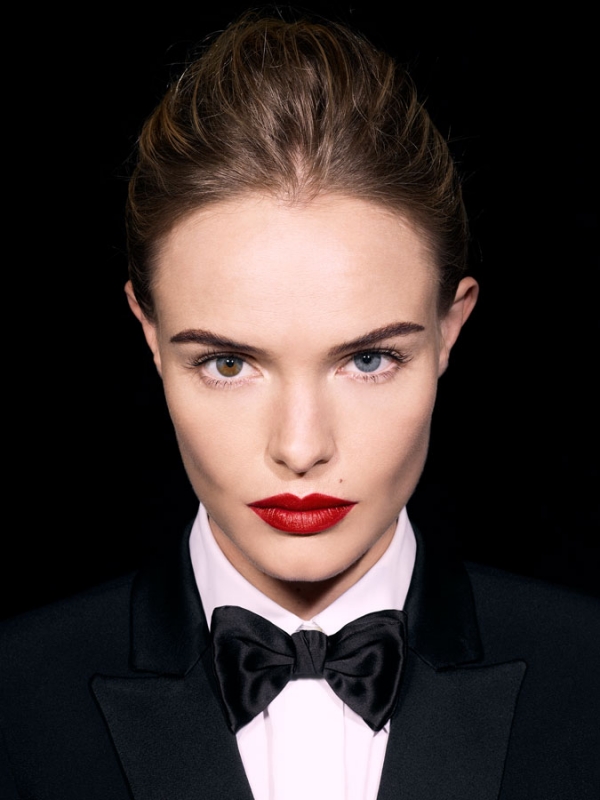 Close-up portrait of a woman in a black tuxedo with a bow tie against a black background with heterochromia and red lipstick.