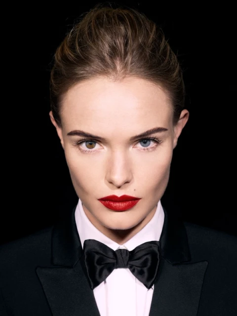 Close-up portrait of a woman in a black tuxedo with a bow tie against a black background with heterochromia and red lipstick.