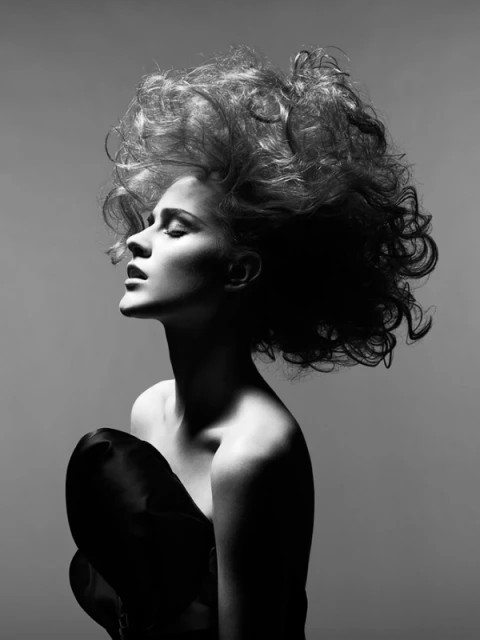 Black and white portrait of a woman with curly hair styled upwards, posed against a gray backdrop wearing a strapless top.